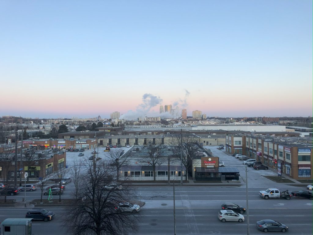 寒々しい郊外都市の夕暮れの風景。高層マンション群を背景に、工場から白い煙があがっている。手前には車が行きかう大通りがある。